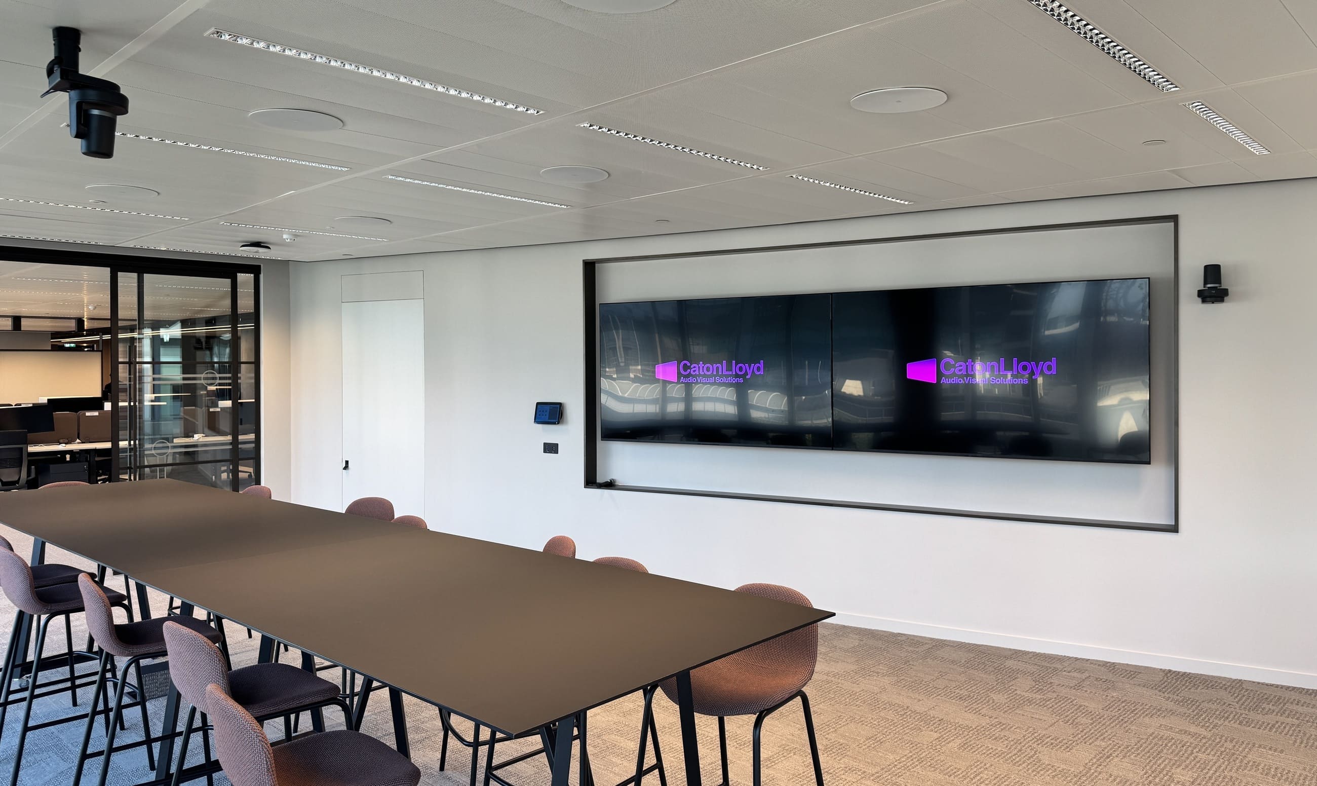collaboration room with twin screens, ceiling and wall mounted cameras, a wall mounted control interface, ceiling mounted microphones and a large table in the centre of the room